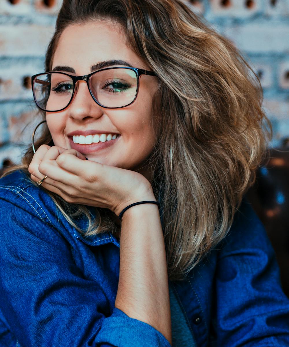 A person wearing glasses and a denim shirt is smiling with their chin resting on their hand in front of a brick wall.