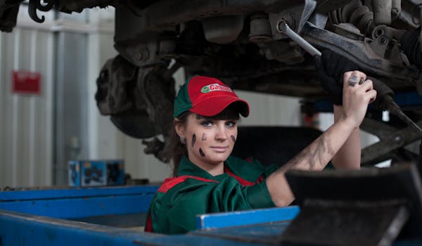 A mechanic in a red and green uniform is working on a vehicle. They have smudges on their face and arms.