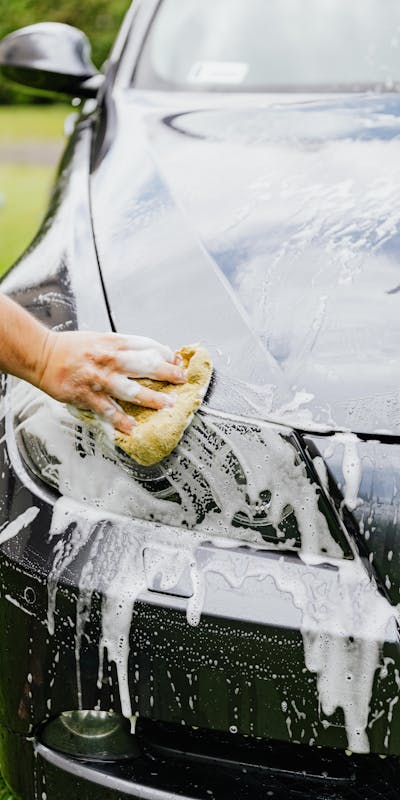 A hand washing a black car's hood with a sponge, covered in soapy suds.