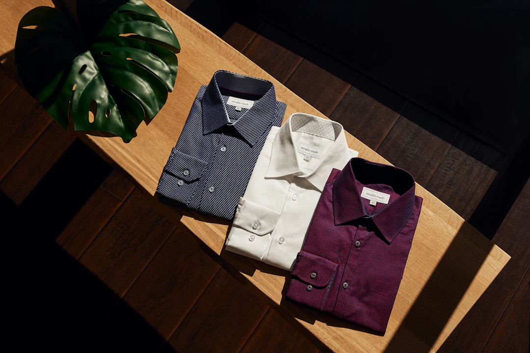 Three neatly folded dress shirts in navy, white, and burgundy displayed on a wooden table, alongside a green leaf for a stylish presentation. Bath Digital Marketing Agency