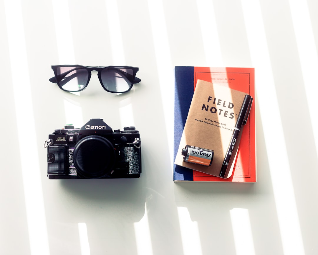A flat lay of a Canon camera, sunglasses, a notebook titled "FIELD NOTES," and a pen, arranged on a surface with natural light casting striped shadows. Bath Digital Marketing Agency