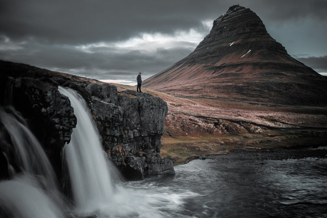 A person stands on the edge of a rocky cliff near a waterfall, overlooking a dramatic landscape with a prominent mountain in the background. The scene is moody, with overcast skies enhancing the natural beauty. Bath Digital Marketing Agency