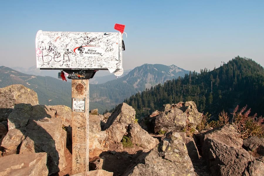 A decorated mailbox on a rocky outcrop, overlooking a mountainous landscape. The mailbox is covered in signatures and messages, with trees and rugged hills in the background Bath Digital Marketing Agency