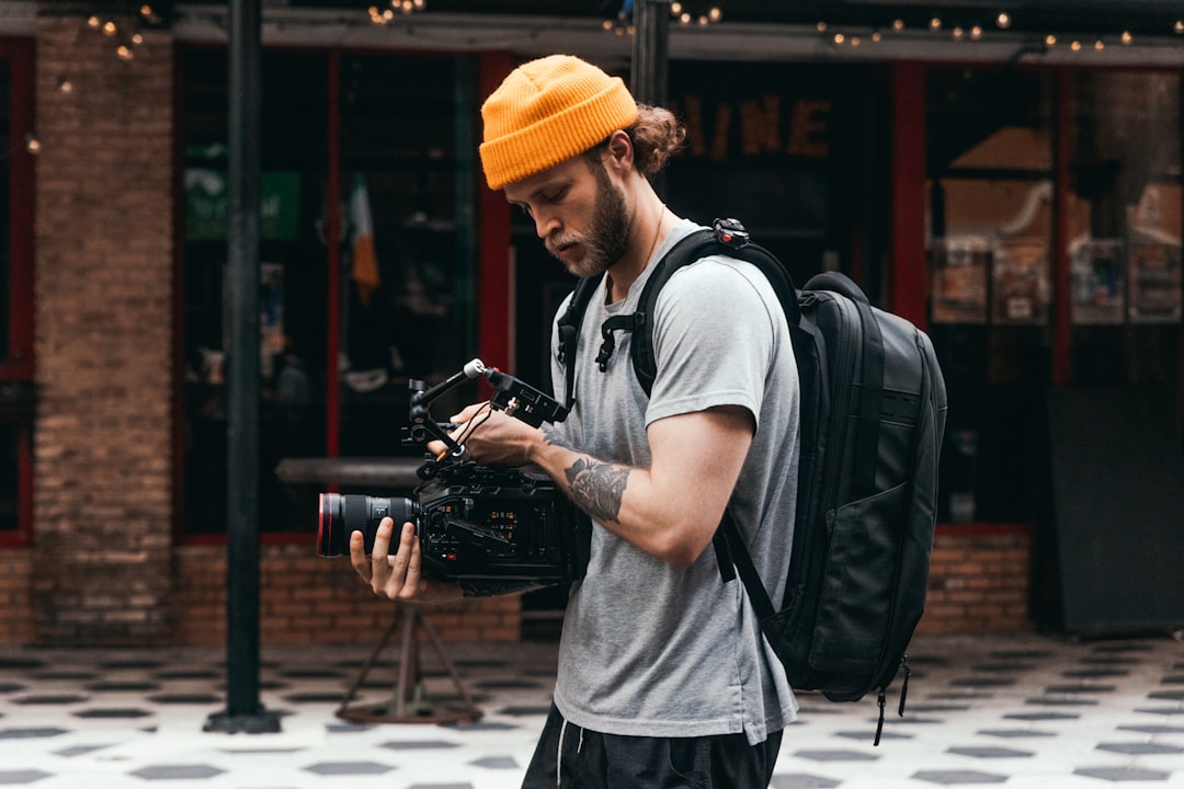 A man wearing a yellow beanie and a grey shirt, examining a camera while carrying a backpack, set against an urban backdrop. Bath Digital Marketing Agency