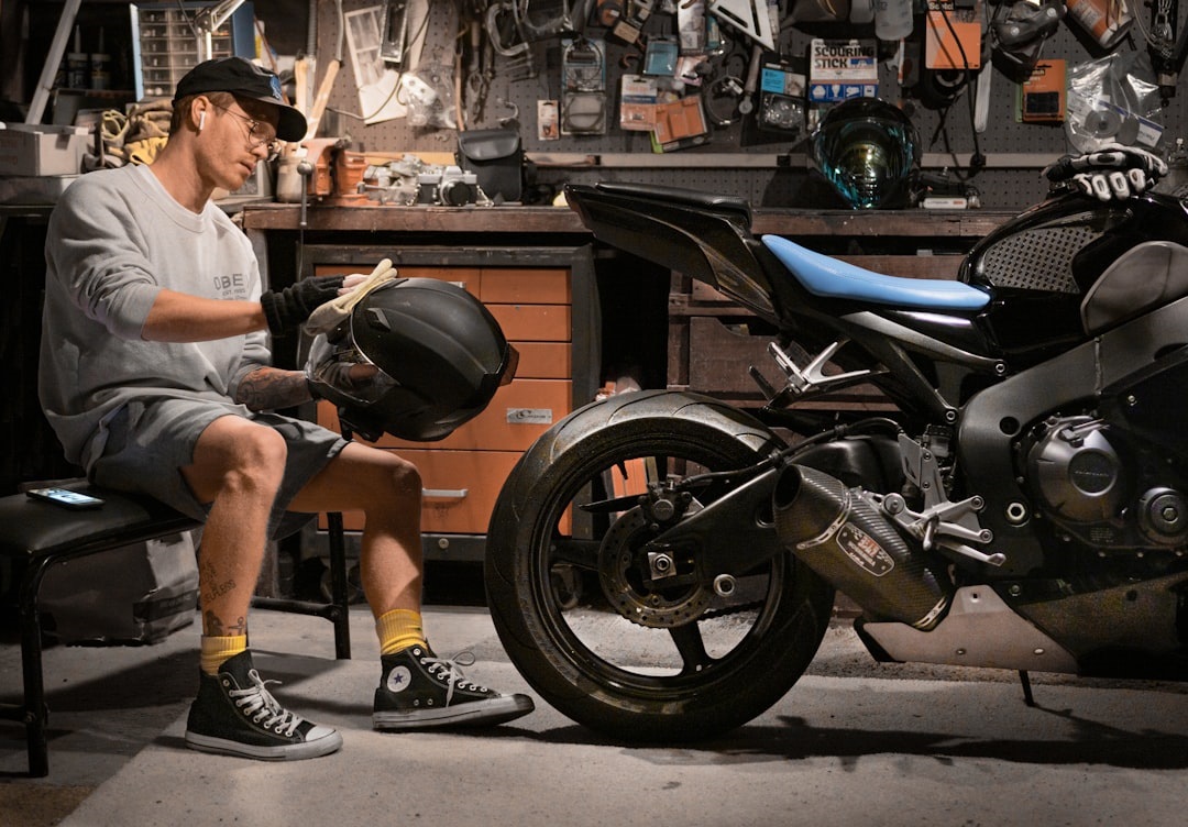 A man in casual attire sitting on a bench, inspecting a motorcycle helmet while a sleek motorcycle is parked beside him in a workshop filled with tools and equipment Bath Digital Marketing Agency