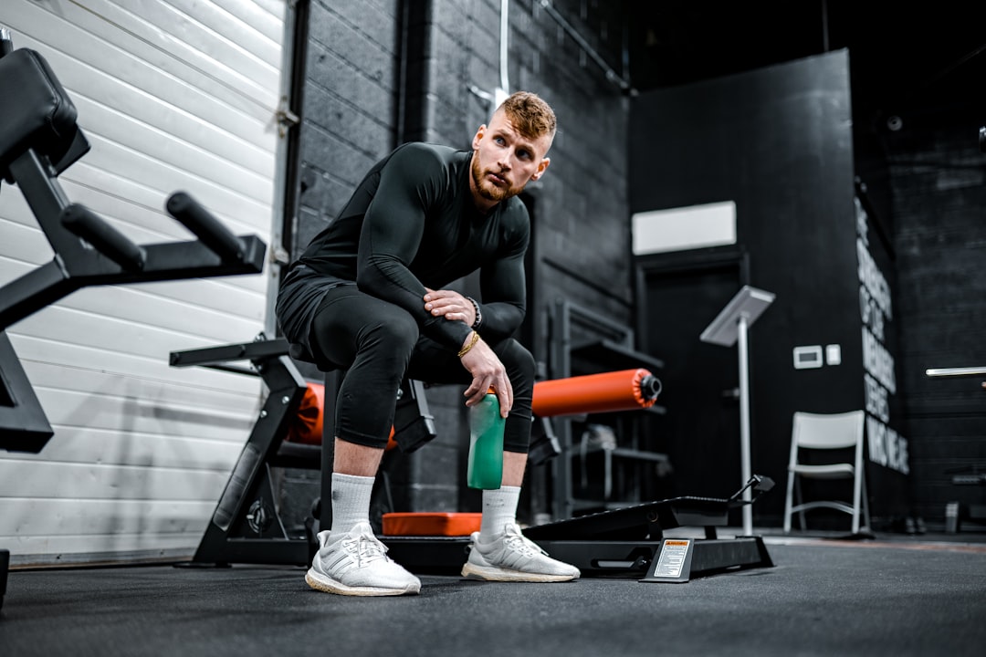 A man sitting on a gym bench, wearing a black long-sleeve shirt and black pants, holding a green water bottle in a fitness studio. Bath Digital Marketing Agency