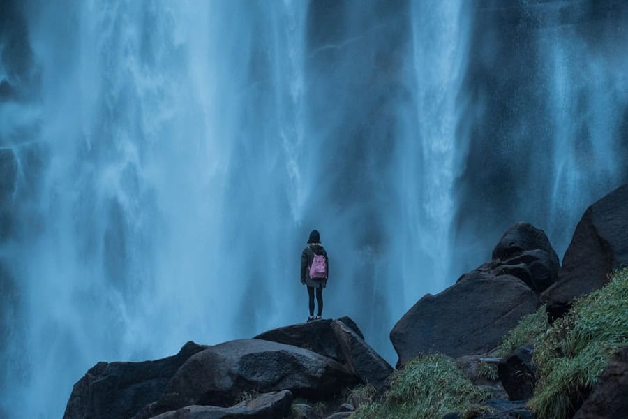 A person standing on a rock in front of a large waterfall, wearing a backpack. The scene captures the powerful flow of water and the surrounding natural landscape. Bath Digital Marketing Agency
