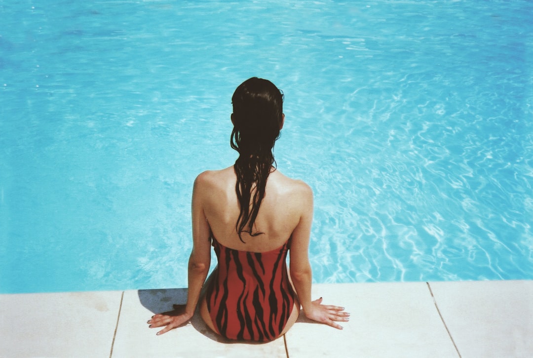 A woman sitting by the edge of a swimming pool, wearing a red and black striped swimsuit, with her back to the camera and water reflecting in the background. Bath Digital Marketing Agency
