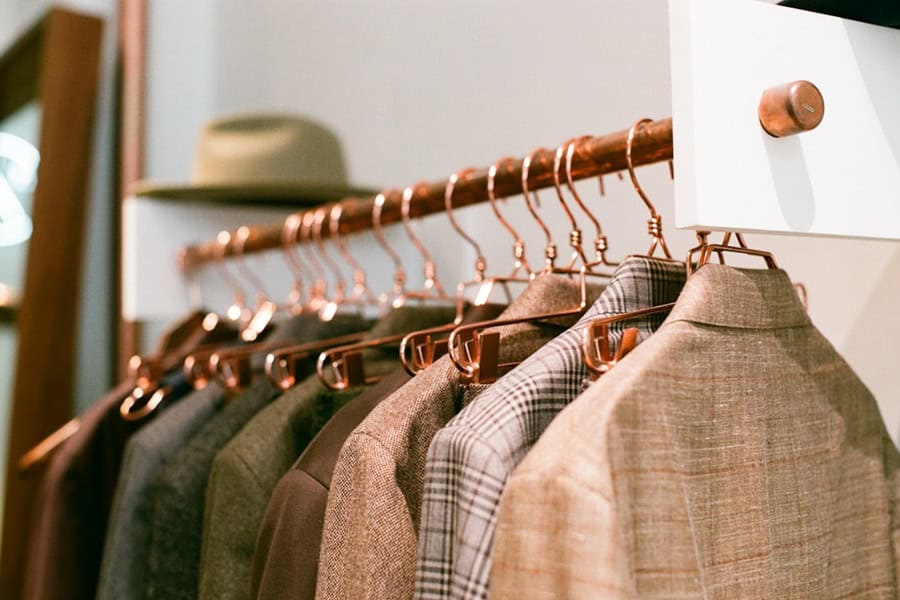 A monochrome image of a rack displaying various suits and jackets, with a hat hanging above. The garments are neatly arranged on hangers, showcasing different textures and patterns. Bath Digital Marketing Agency