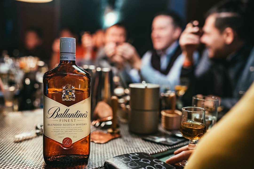 A bottle of Ballantine's Finest Blended Scotch Whisky prominently displayed on a bar table, with blurred figures of people enjoying drinks in the background. Bath Digital Marketing Agency