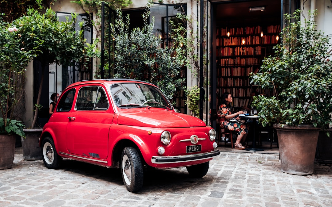 A vintage red car parked outside a quaint café surrounded by greenery, with a person sitting inside the café in the background. Bath Digital Marketing Agency