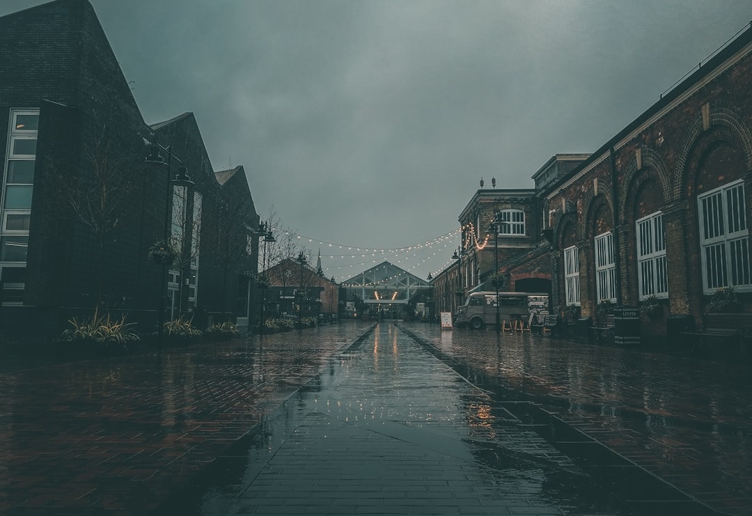 A moody, rainy street scene featuring brick buildings and string lights. The wet pavement reflects the overcast sky, creating a tranquil yet atmospheric setting.  Bath Digital Marketing Agency