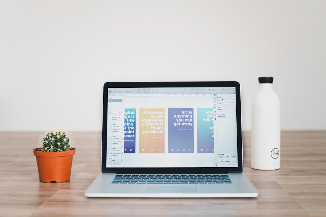 A laptop displaying a graphic design project, accompanied by a potted cactus and a water bottle on a wooden desk against a plain background. Bath Digital Marketing Agency