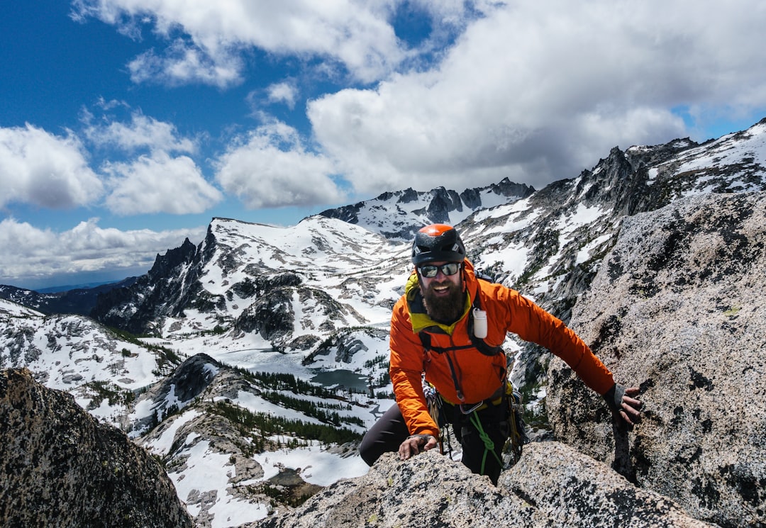 A climber in an orange jacket and helmet poses on a rocky mountain peak, with snow-capped mountains and a blue sky in the background. Bath Digital Marketing Agency