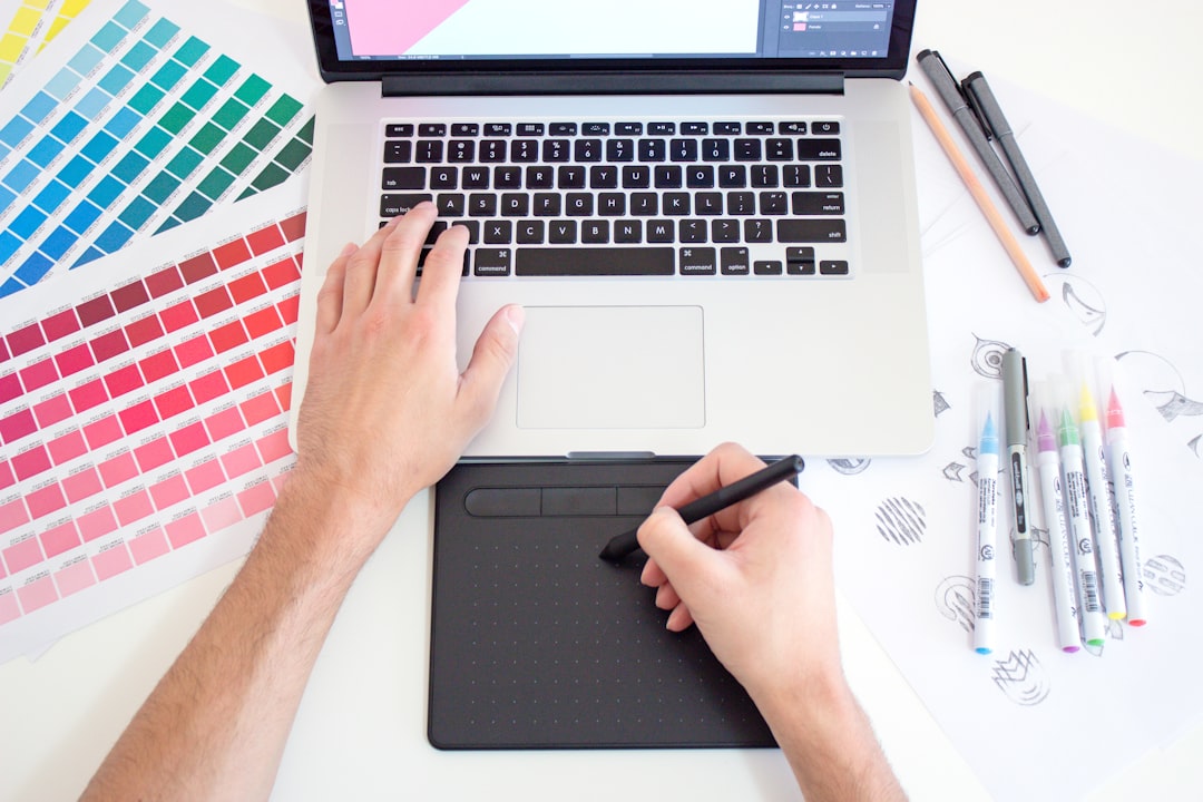 A person's hand using a stylus on a graphic tablet beside a laptop, surrounded by colour swatches and various drawing pens on a white desk. Bath Digital Marketing Agency