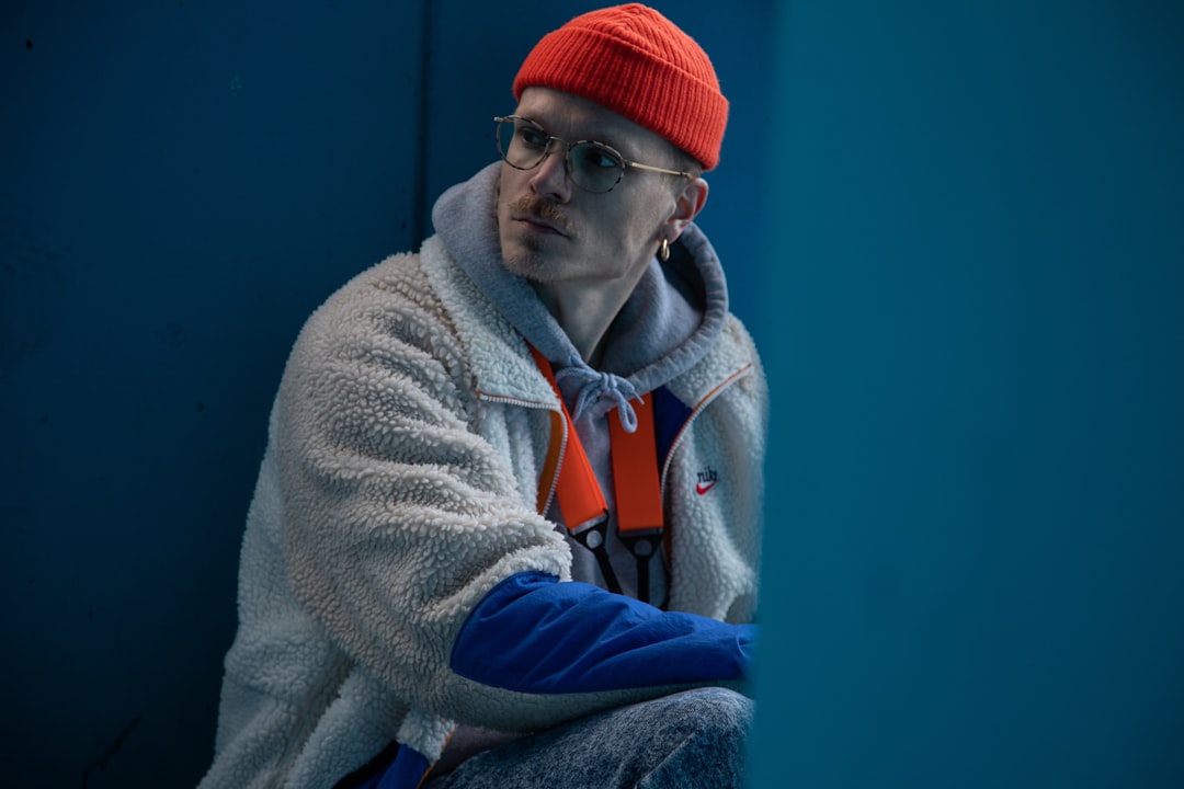 A young man wearing glasses and a bright orange beanie sits against a blue background. Bath Digital Marketing Agency