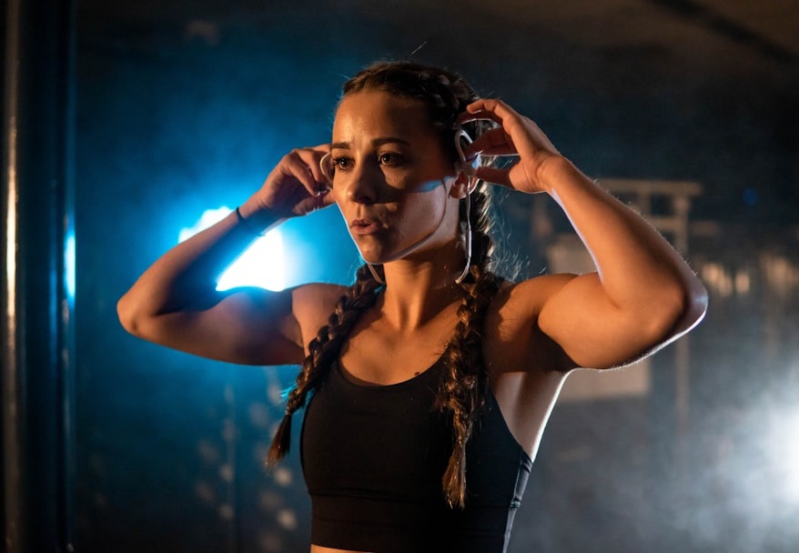 A fit woman adjusts her headphones while preparing for a workout. She has braided hair and is illuminated by dramatic lighting, highlighting her focused expression and athletic physique. Bath Digital Marketing Agency
