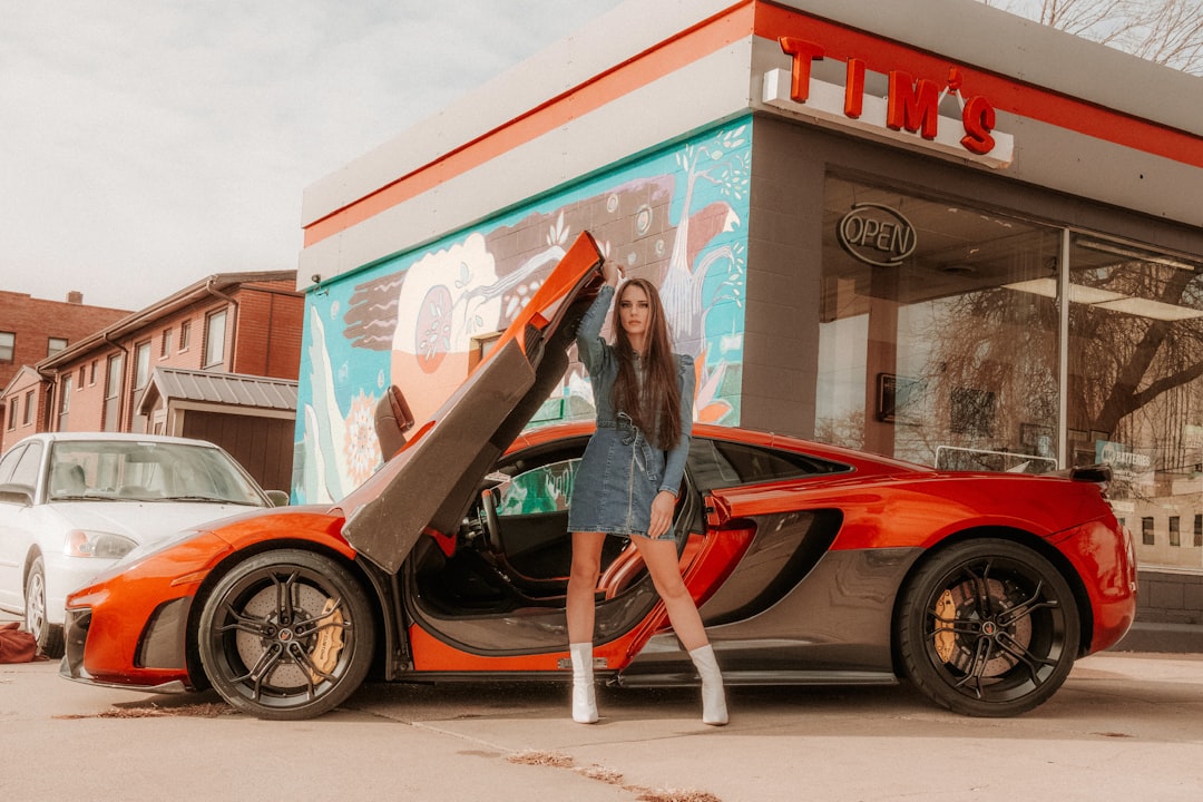 wo bs on display in tA woman stands beside an orange McLaren with its door open, posing confidently in front of a brightly painted café. She wears stylish silver boots and a denim outfit, complementing the vibrant setting. Bath Digital Marketing Agency