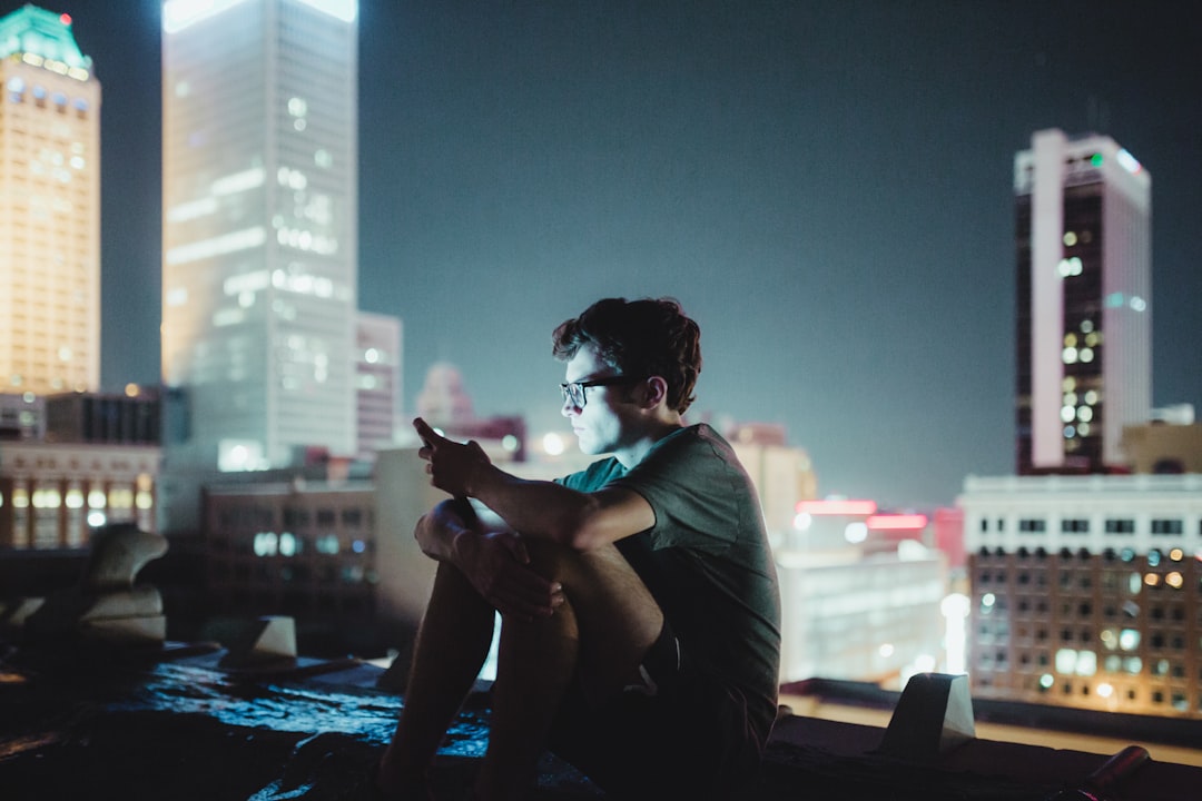 A young man sits on a rooftop at night, illuminated by the light of his phone. The city skyline with tall buildings glimmers in the background, creating a vibrant urban scene. Bath Digital Marketing Agency