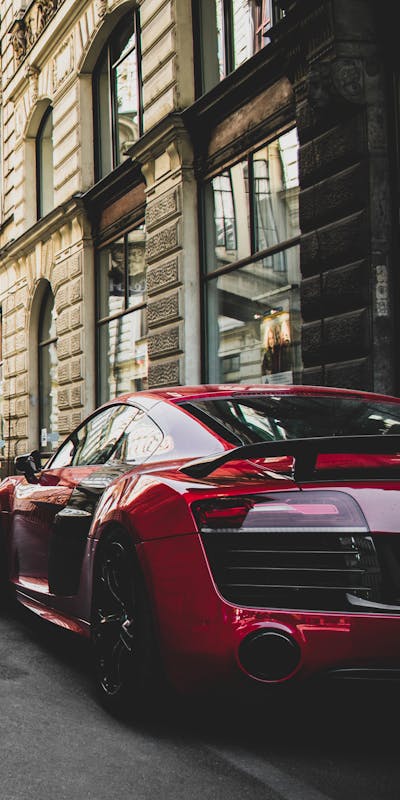 A red sports car parked on a city street, viewed from the rear, near a building with large windows.