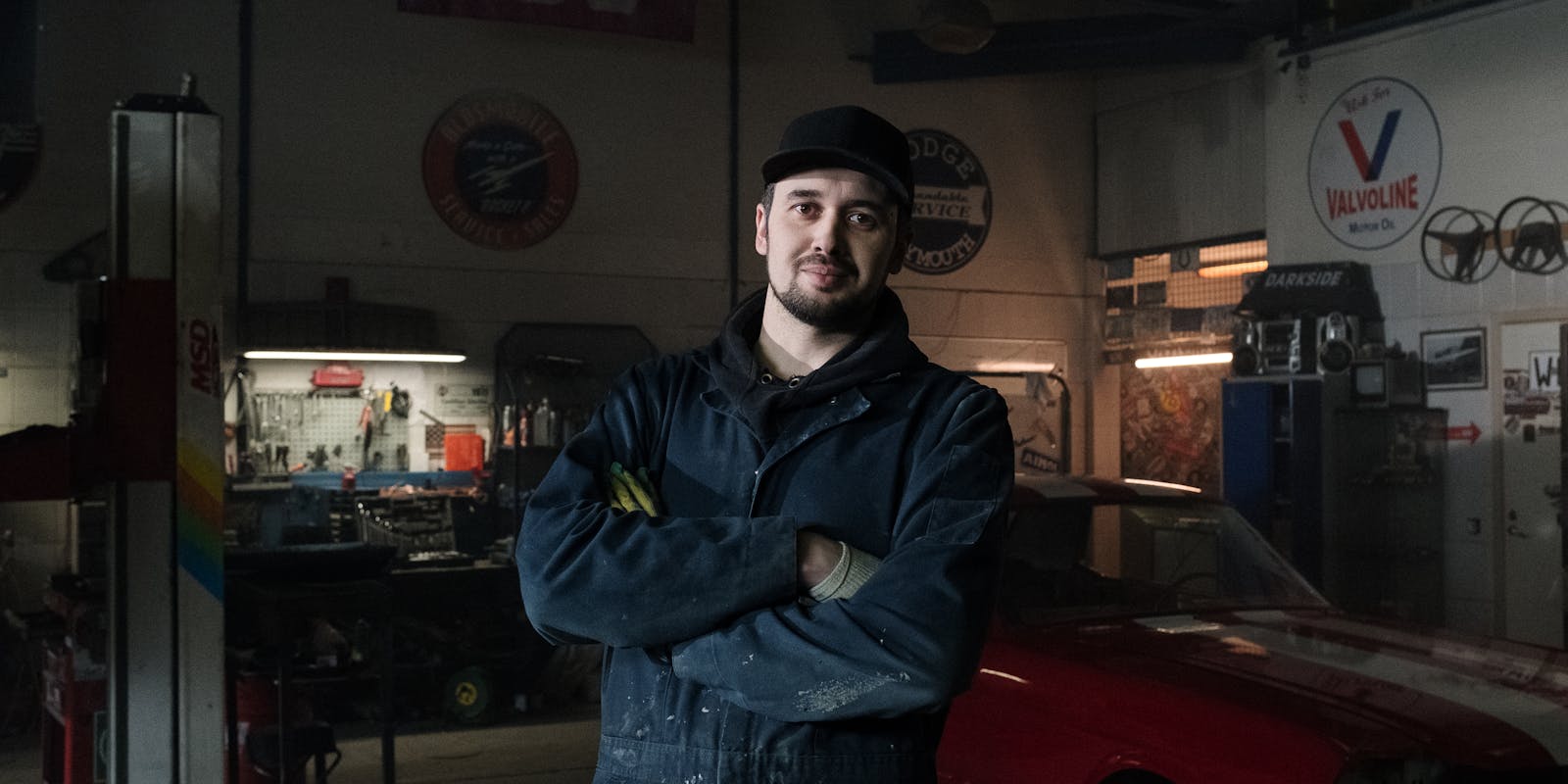 A man in a dark mechanic's jumpsuit and cap stands with arms crossed in a dimly lit garage, surrounded by tools and car parts.