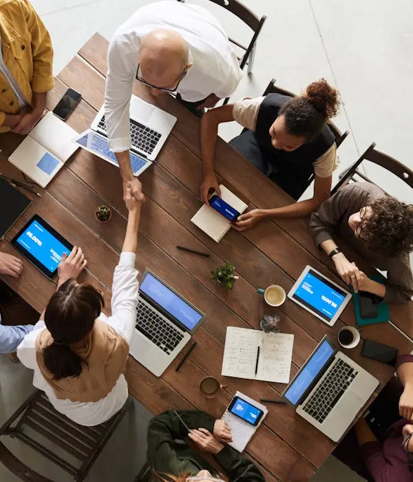 Overhead view of a diverse group of colleagues working together at a wooden table, with laptops, tablets, notebooks, and coffee cups. Two team members are shaking hands, indicating collaboration or agreement
