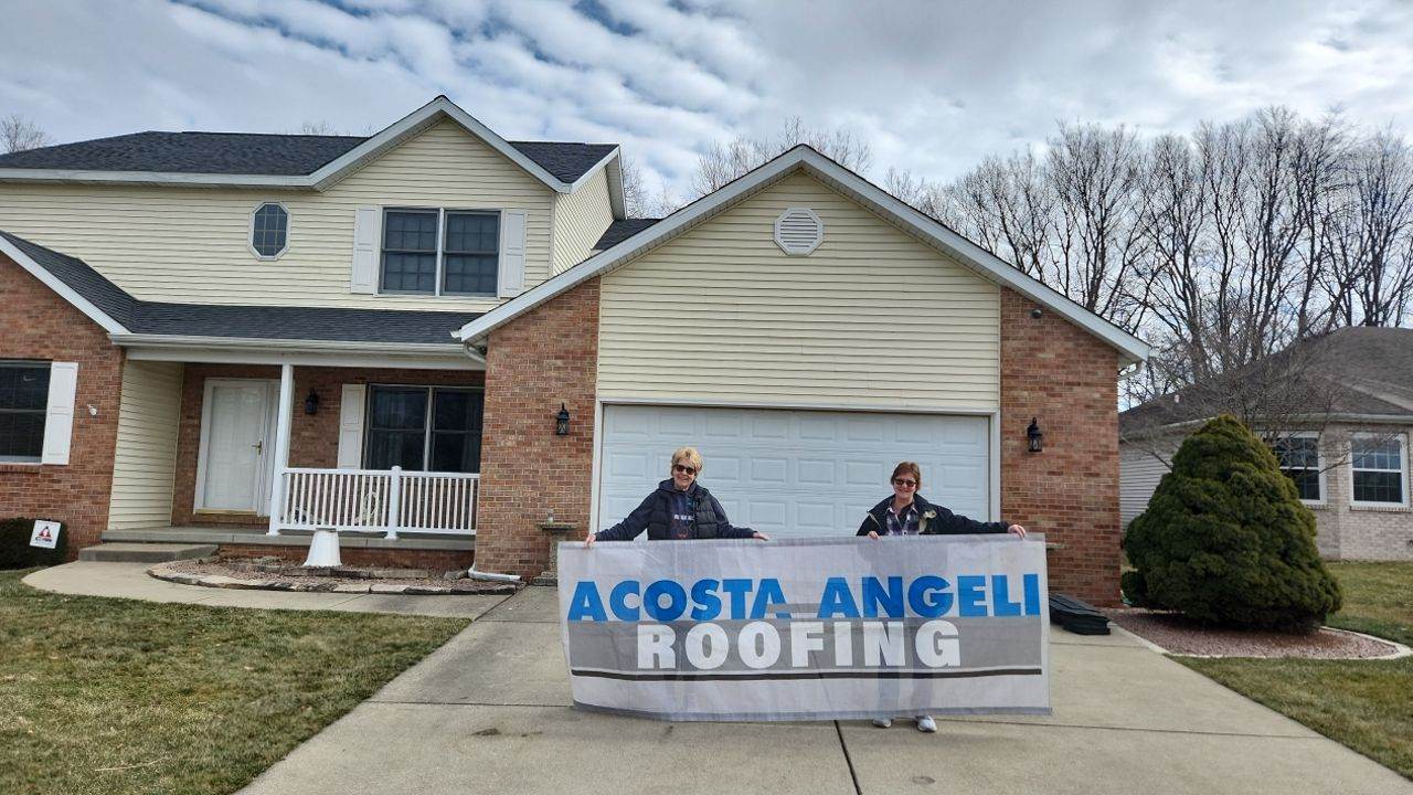 Two individuals holding a "Acosta-Angeli Roofing" banner in front of a residential house.