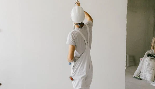 A man wearing a white hard hat is painting a wall with a paint roller.
