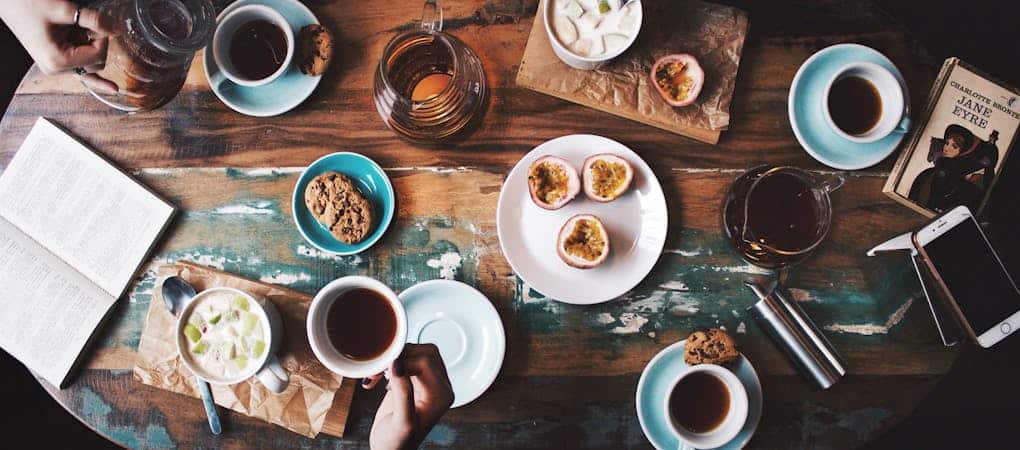 Overhead view of a rustic wooden table with several cups of coffee and tea, cookies, passion fruit halves, open books, and smartphones - beann speciality coffee