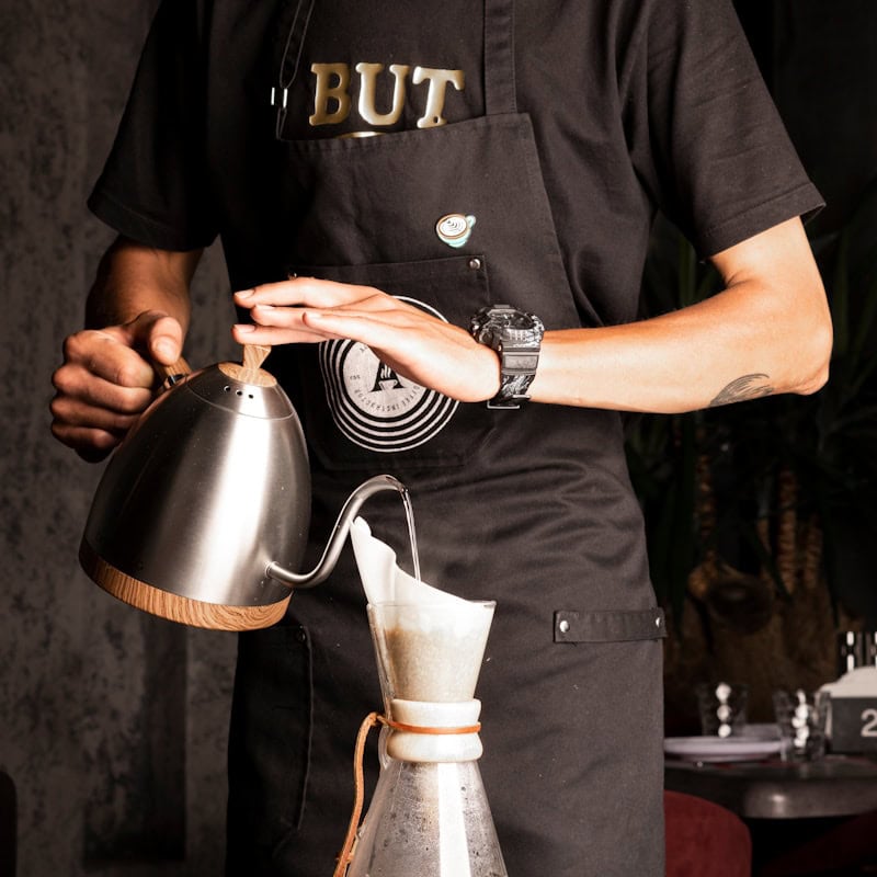 A person in an apron pouring water from a gooseneck kettle into a pour-over coffee maker - beann artisan coffee