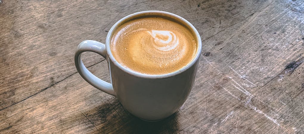 A white mug filled with a cappuccino featuring a heart-shaped latte art design, sitting on a rustic wooden table - beann artisan coffee