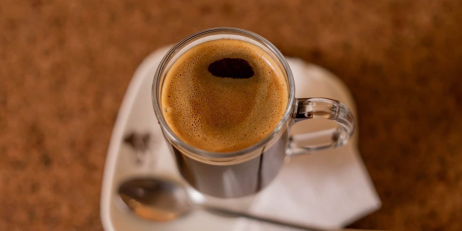 Top-down view of a glass mug filled with Beann artisan coffee, featuring a thick layer of crema, on a white saucer with a spoon -  beann speciality coffee