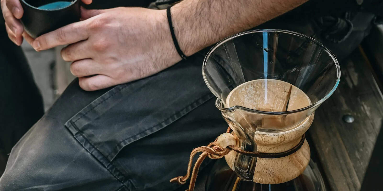 Person holding a small cup next to a glass pour-over coffee maker with a wooden collar - beann artisan coffee