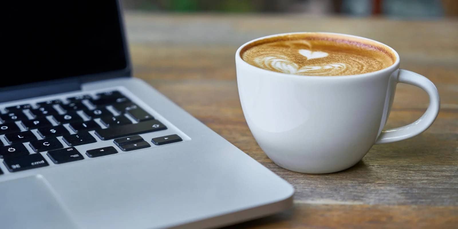 A white cup of coffee with latte art next to a silver laptop on a wooden table - beann artisan coffee