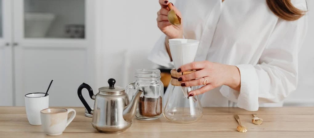 A woman in a white shirt making artisan pour-over coffee at home using a glass coffee maker and a gold spoon - beann artisan coffee