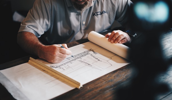 A man sits at a table, focused on a blueprint, with drafting tools and papers scattered around him.