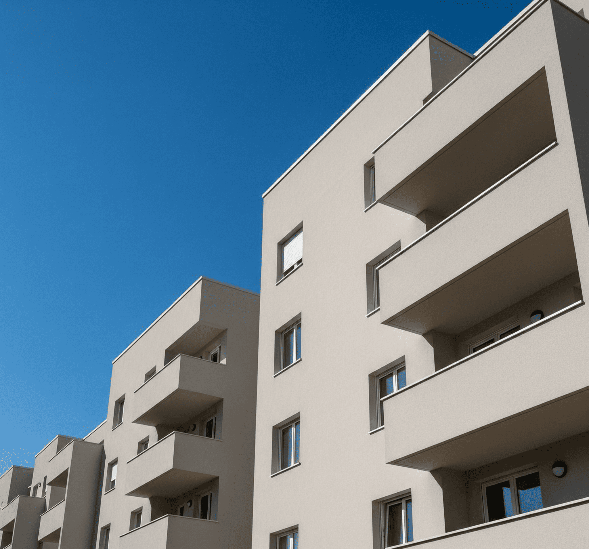 A modern, light beige stucco apartment complex with large balconies is viewed from below against a vivid blue sky.