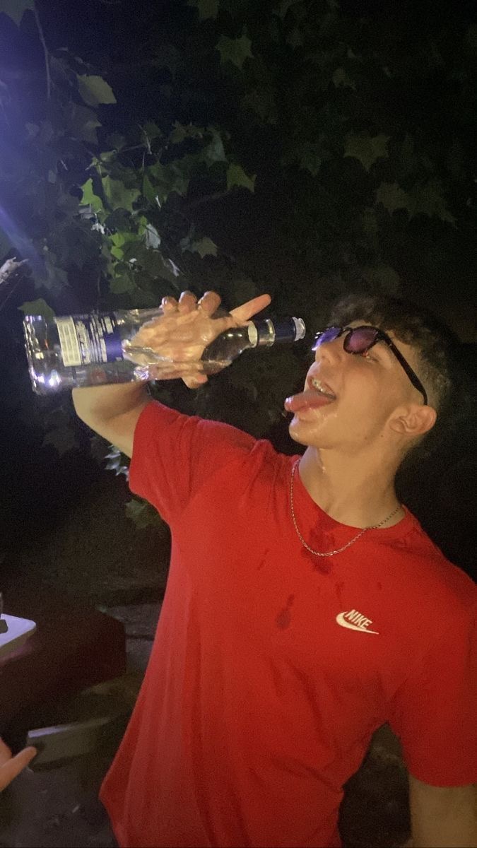 Personne portant un t-shirt rouge, buvant directement à la bouteille dans un cadre extérieur sombre, avec une ambiance festive.