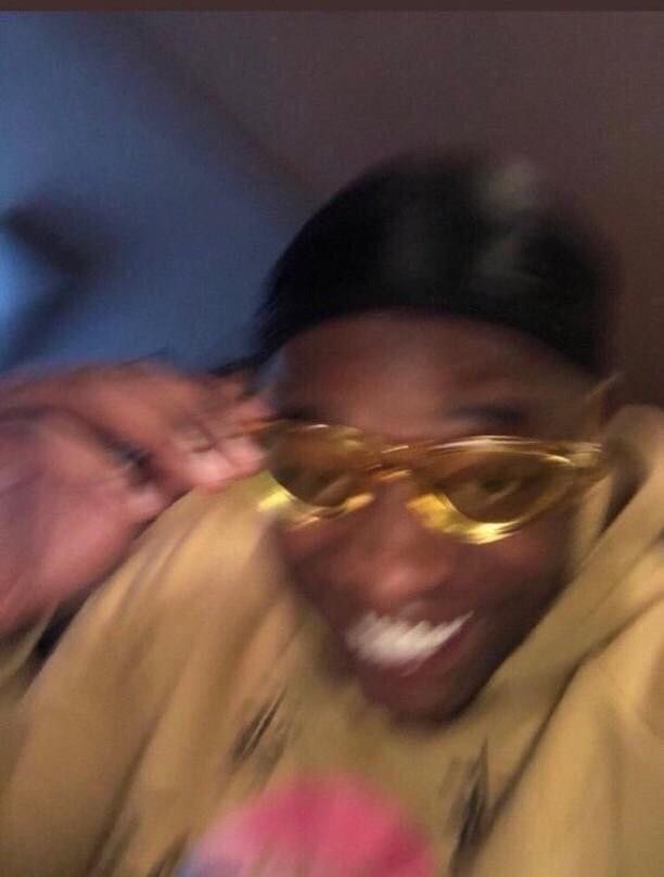 Un homme souriant portant des lunettes de soleil dorées et un sweat à capuche jaune, capturé dans une photo légèrement floue, ajoutant une touche énergique et amusante