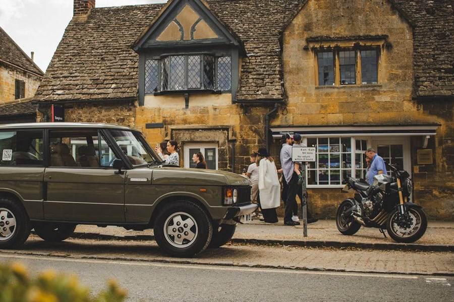 A vintage olive green Range Rover Classic is parked on a street in a village with old stone buildings, with several people standing nearby and a black motorcycle parked on the sidewalk | Castle Combe England