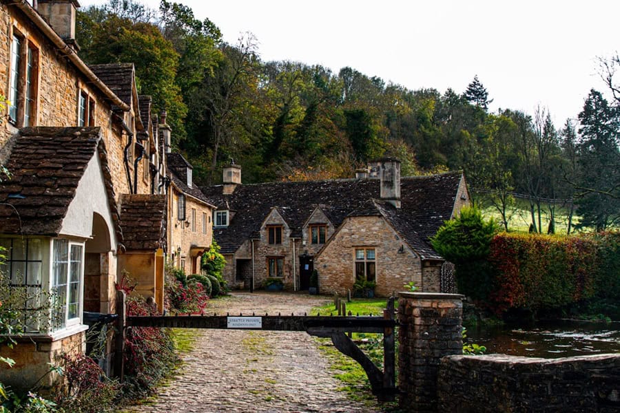 A view of a quaint village street in the Cotswolds, England, featuring historic, honey-coloured stone cottages and a small wooden bridge over a stream | Castle Combe England
