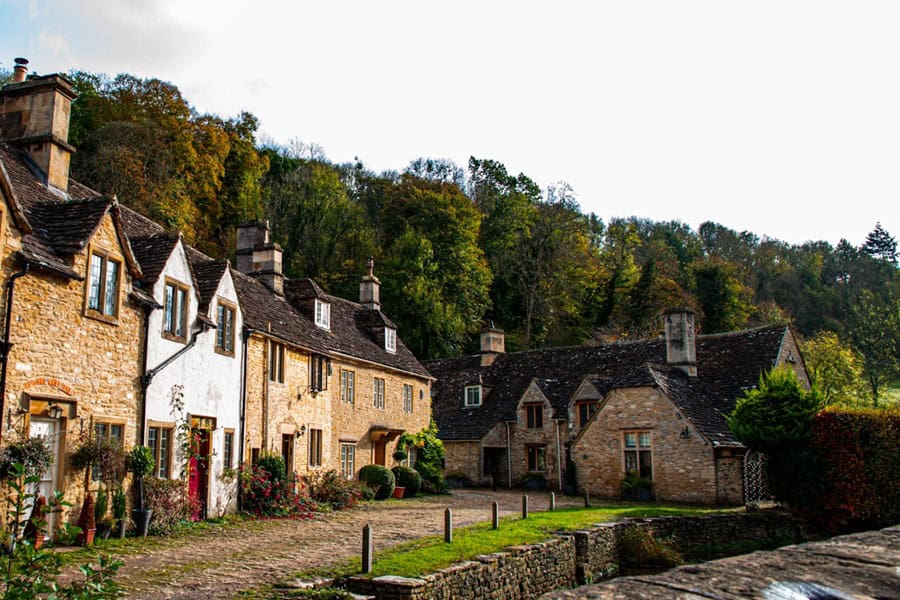 A row of historic, stone weavers' cottages in the village of Castle Combe, England, backed by a lush, green hill | Castle Combe England