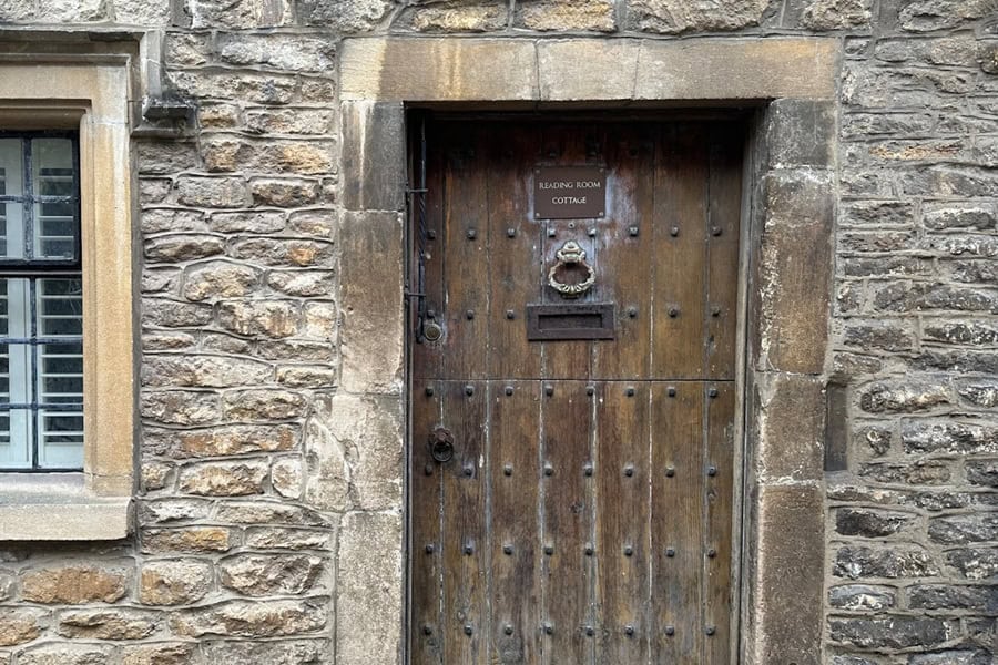 A stone cottage wall featuring a weathered wooden door with a sign reading 'READING ROOM COTTAGE' | Castle Combe England