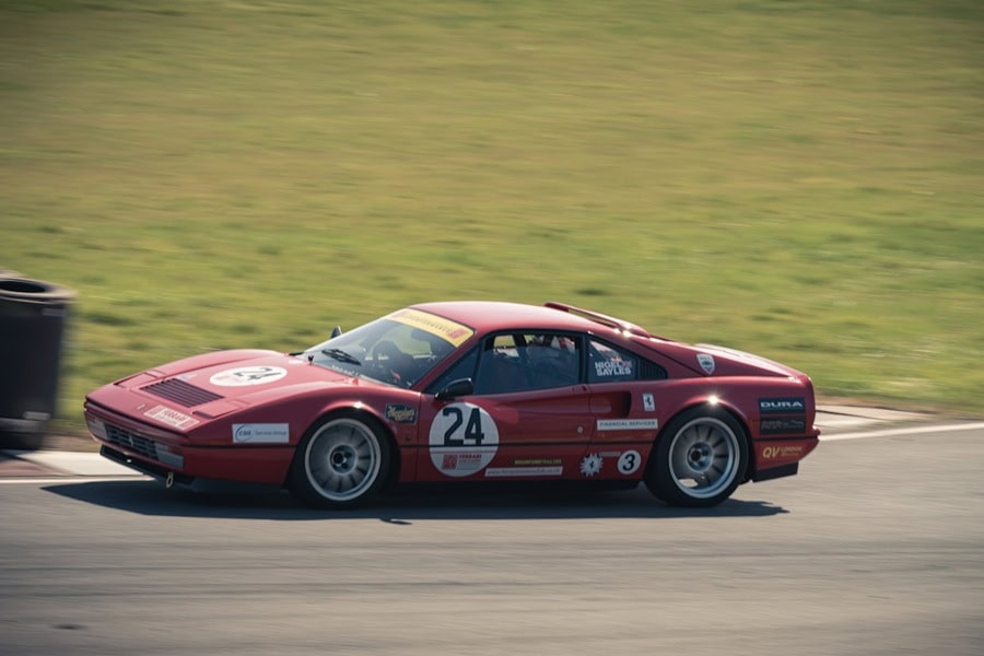 A red Ferrari 328 GTB race car with the number 24 on its side speeds around a racetrack during a classic car race event | Castle Combe England