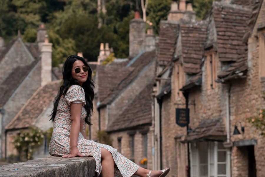 A woman in a floral dress and sunglasses sits on a stone wall overlooking the picturesque stone cottages in the English village of Castle Combe | Castle Combe England