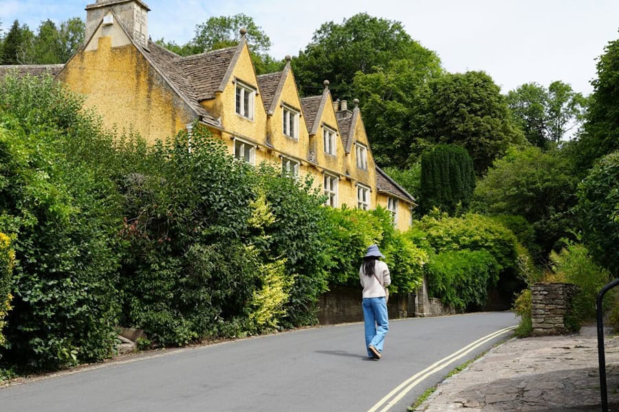 A person walks down a quiet asphalt road in Castle Combe, a picturesque village in the Cotswolds, England | Castle Combe England