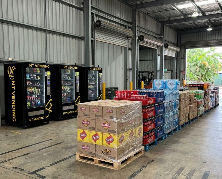 A view inside a warehouse shows three black vending machines with yellow "NT VENDING" branding lined up against a metal wall on the left. In the center and extending to the right, several wooden pallets are stacked with products, including boxes of Smith's Chips and Twisties, cases of Coca-Cola and Pepsi, and shrink-wrapped packs of water bottles. Large industrial roller doors are open in the background, revealing tropical plants outside. The warehouse has a concrete floor and a high metal ceiling.