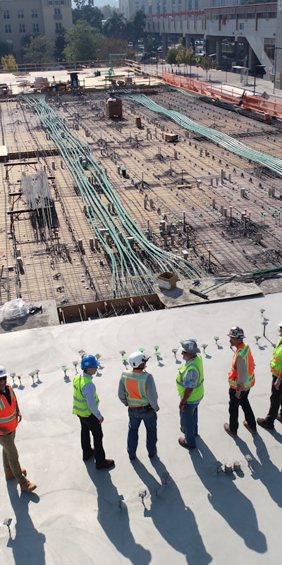 A group of construction workers in hard hats stands on a construction site, surrounded by equipment and building materials.