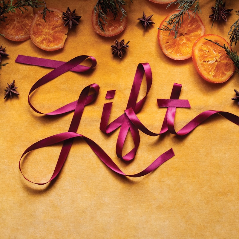 Festive display featuring a luxurious magenta ribbon spelling out the word "gift" in a cursive script, surrounded by dried orange slices and star anise spices. This image represents the artisan gift options available from Bonsaii. 