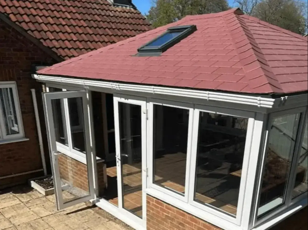Conservatory warm roof conversion featuring terracotta red lightweight shingles, white uPVC windows, and open French doors by Norfolk Exterior Solutions.